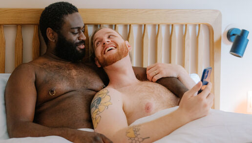 A queer couple uses a phone in bed together (Source: Building Healthy Online Communities Photo Collective)