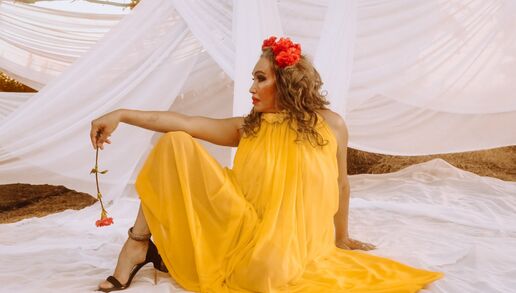 Cecilia Gentili poses in profile wearing a flowing yellow dress and crown of red flowers. She faces left and holds a matching flower in her outstretched left hand, which is balanced on her raised knee. She sits on a white cloth laid on the grass and is surrounded by trees draped in matching white fabric. Sunlight peeks through the trees.