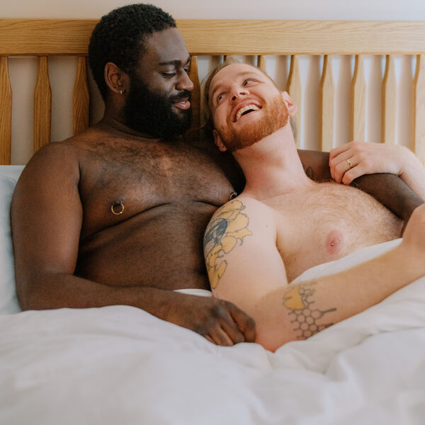 A queer couple uses a phone in bed together (Source: Building Healthy Online Communities Photo Collective)