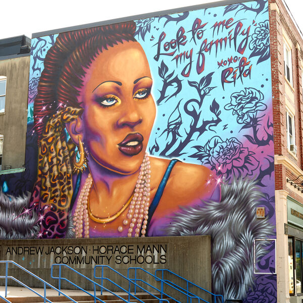 Photo of the mural "Rita's Spotlight" by the artist Rixy. Rita Hester is shown from the shoulders up. Her braids are pulled back with a leopard print scarf and she wears a gold necklace, earrings, and multiple strands of pearls. A fur is draped around her shoulders and she is surrounded by flowers on a blue and purple background. Text to the right of her face reads: "Look to me my family, xoxo, Rita"