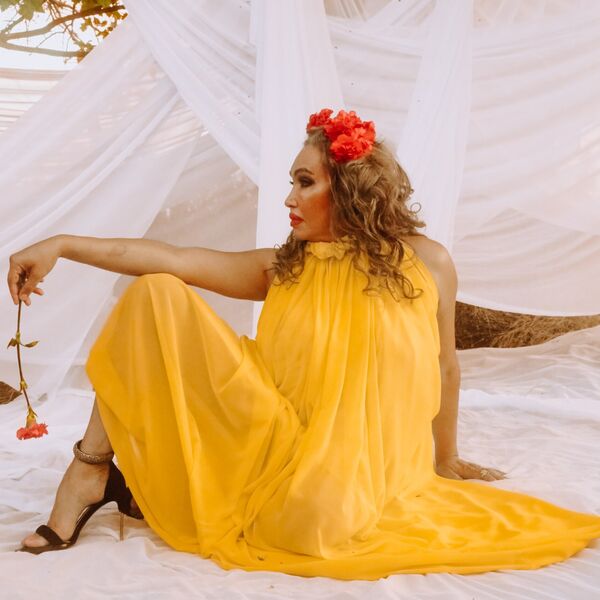 Cecilia Gentili poses in profile wearing a flowing yellow dress and crown of red flowers. She faces left and holds a matching flower in her outstretched left hand, which is balanced on her raised knee. She sits on a white cloth laid on the grass and is surrounded by trees draped in matching white fabric. Sunlight peeks through the trees.